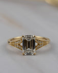 Cora split-shank engagement solitaire featuring an emerald cut diamond accented by round diamonds in a detailed milgrained gold band.