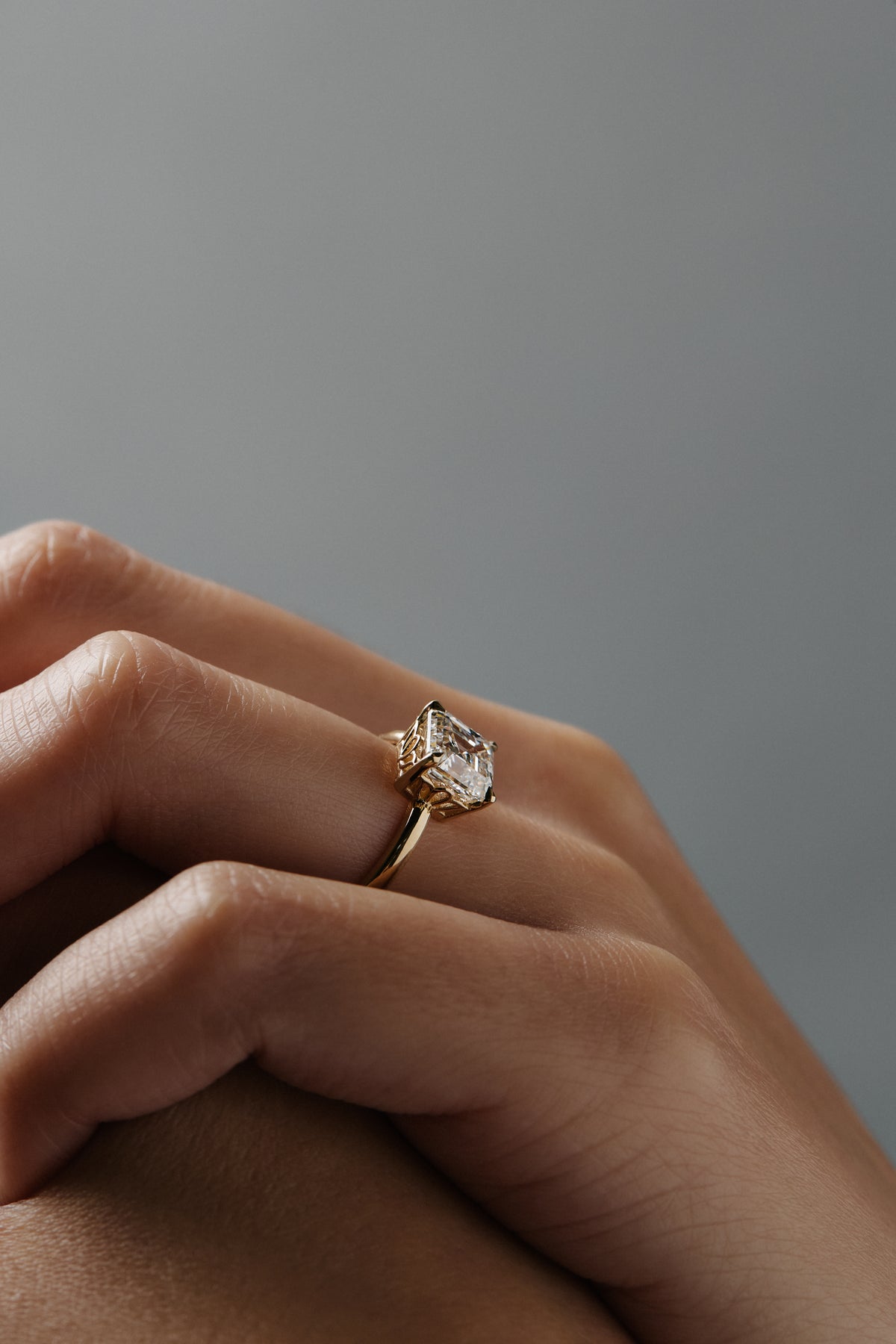 Stylized close-up of Asscher cut diamond claw solitaire worn on a woman's finger, highlighting the delicate floral scroll detailing along the side of the low-profiled setting. 