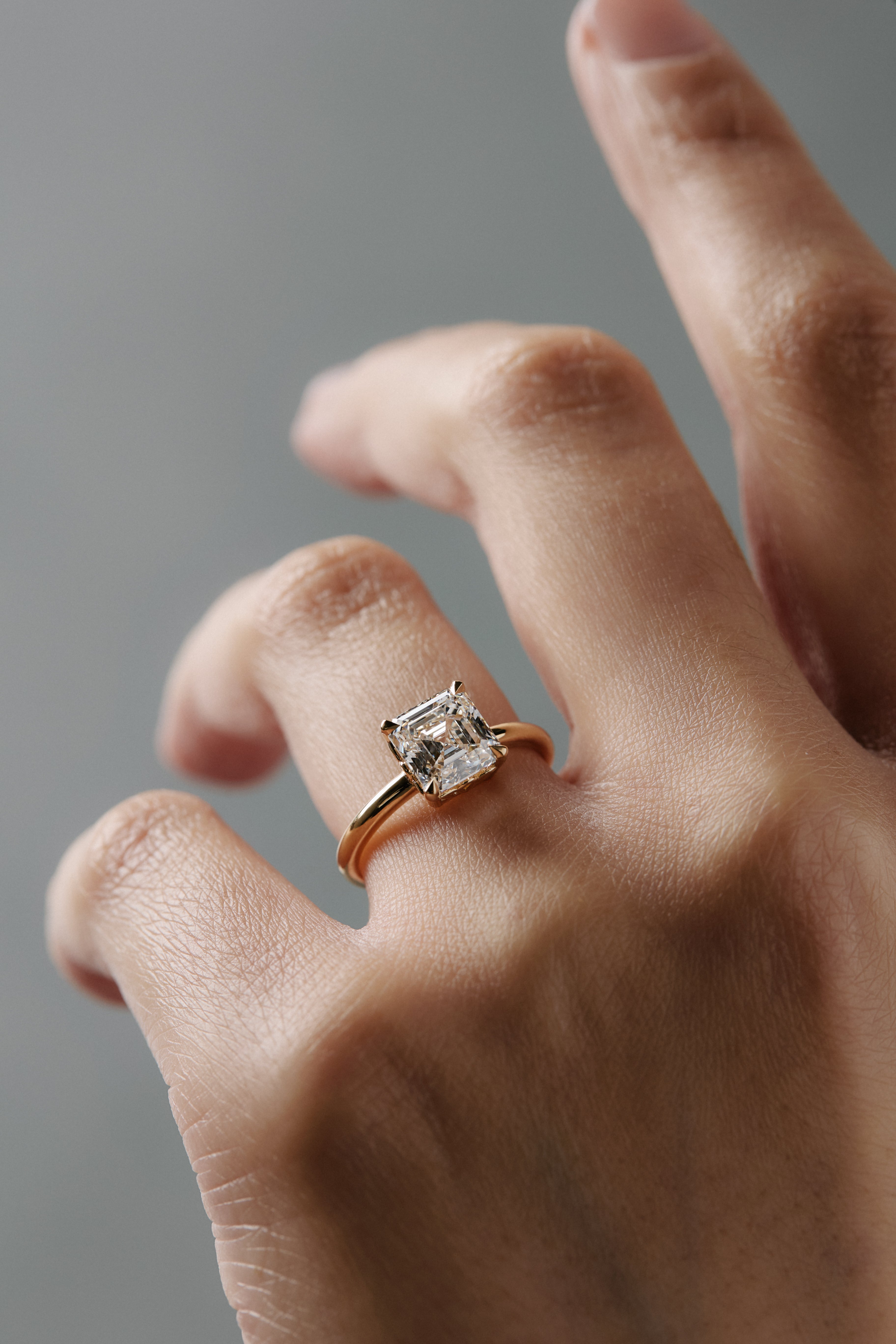Asschercut diamond claw solitaire engagement ring with floral scroll side detailing, worn alone on a woman's finger.