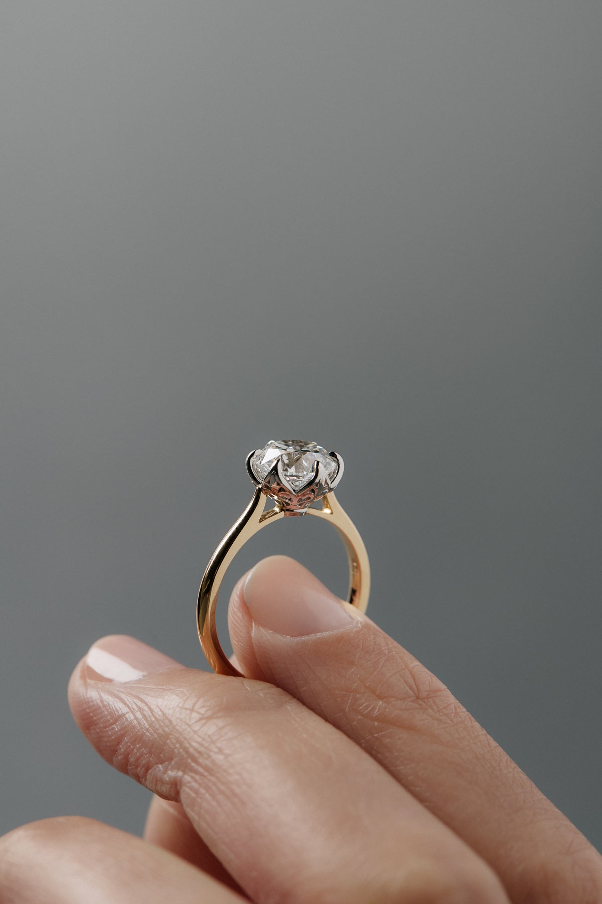 Close-up of the side of the Cala tulip-style cathedral solitaire held between a woman's fingers, showing the antique floral filigree inspired platinum and gold setting. 