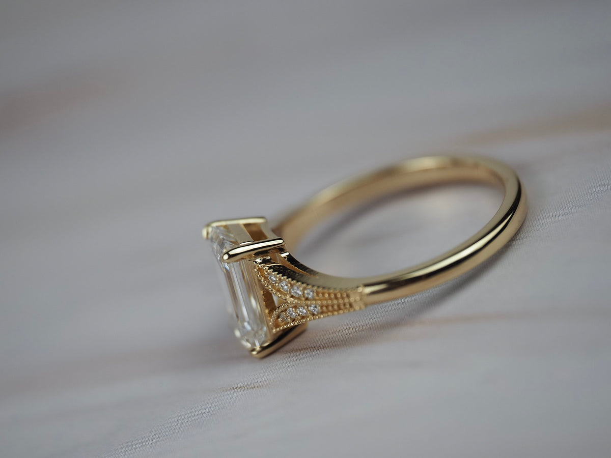 Close-up side profile o father Cora split-shank milgrain engagement solitaire showing the emerald cut diamond, round diamond accents, and detailed gold setting.