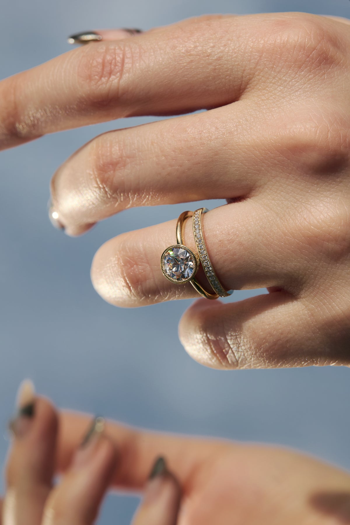 Close-up of a handcrafted milgrain Fairmined gold wedding band paired with the Flora solitaire engagement ring on a woman's hand.